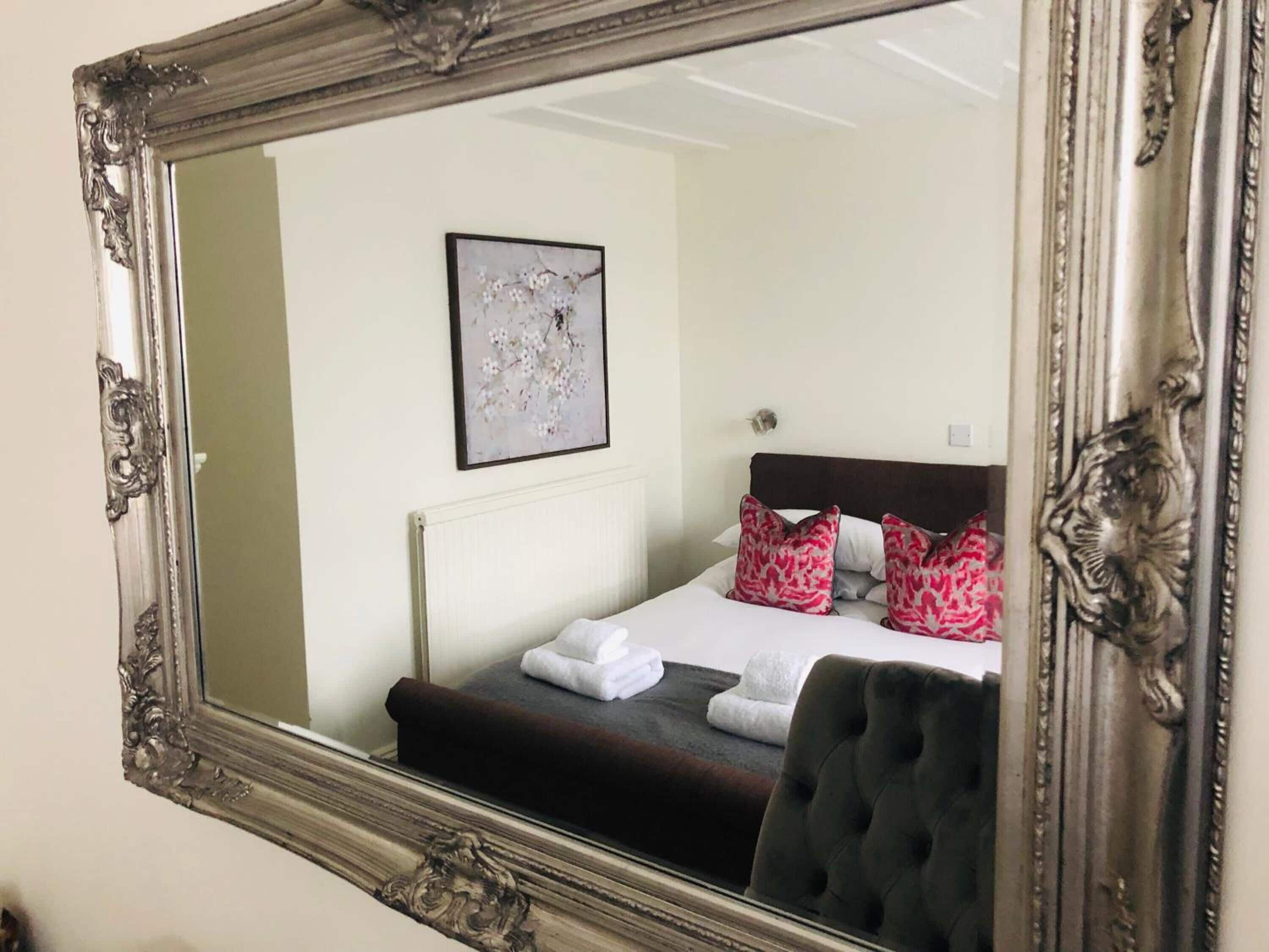 Un dormitorio con un espejo grande y ornamentado, una cama con ropa de cama oscura, dos almohadas, una obra de arte abstracta enmarcada en la pared y una butaca tapizada en tonos oscuros.