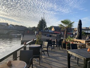 Terraza a la orilla del río con mesas al aire libre, un puente al fondo y una estatua.