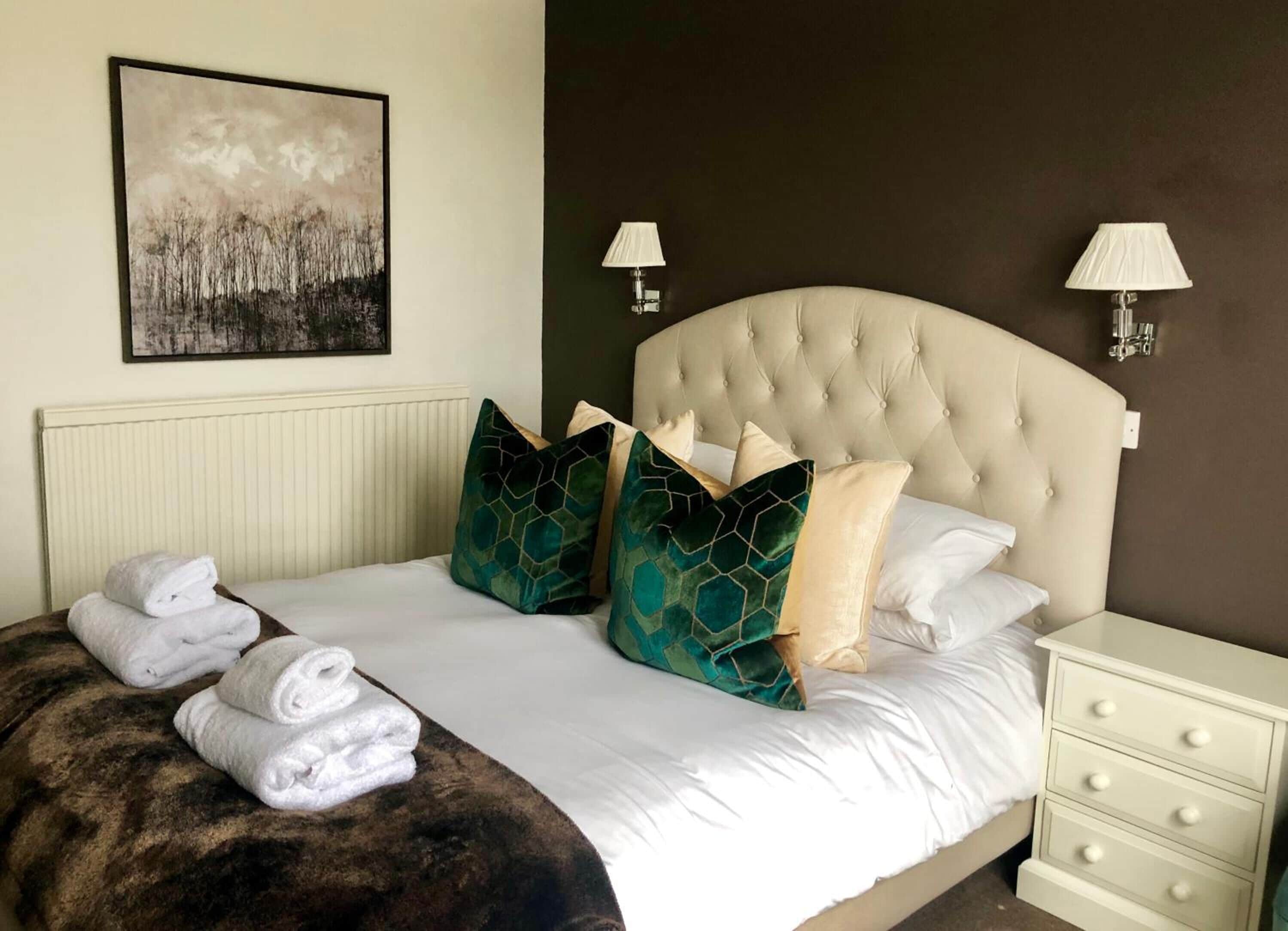 A neatly made bed with a tufted headboard, white linens, and decorative pillows. There are two bedside tables with lamps, a radiator under the bed, and a framed artwork on the wall.
