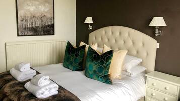 A neatly made bed with a tufted headboard, white linens, and decorative pillows. There are two bedside tables with lamps, a radiator under the bed, and a framed artwork on the wall.