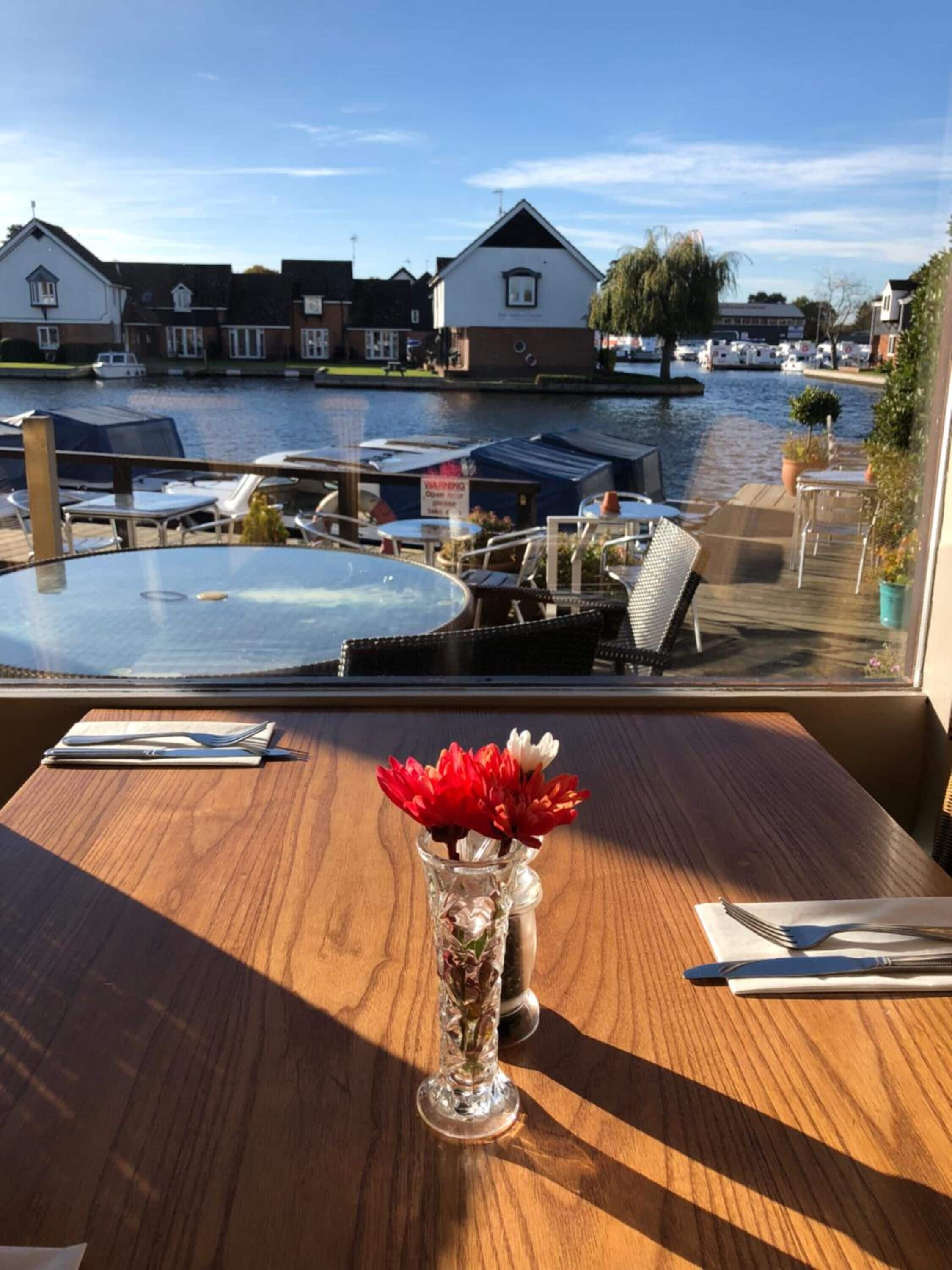 Una mesa de madera con un florero de flores rojas, un menú y cubiertos. Se ve una vista de un canal con casas y barcos a través de una ventana.