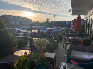 Un patio con mobiliario exterior, vista a barcos en el agua y un atardecer al fondo.