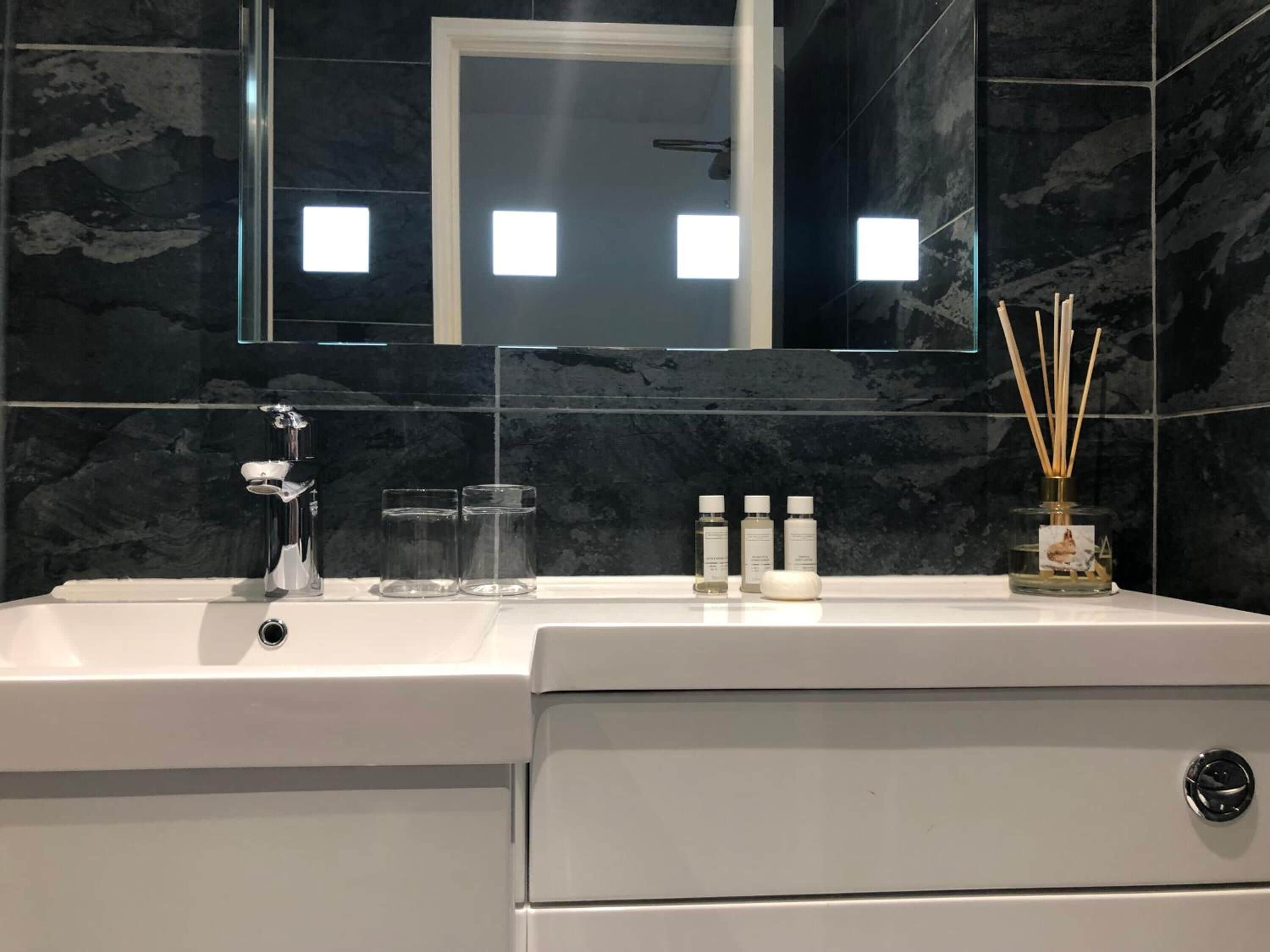 A modern bathroom with a white sink, a mirror, and a marble backsplash. There are three bottles of liquid soap and two glass containers on the sink.