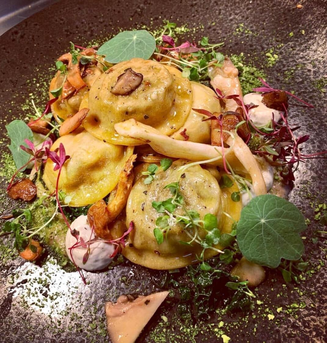 En rätt med ravioli, svamp och grönsaker, serverad på en mörk tallrik eller ett mörkt underlag.