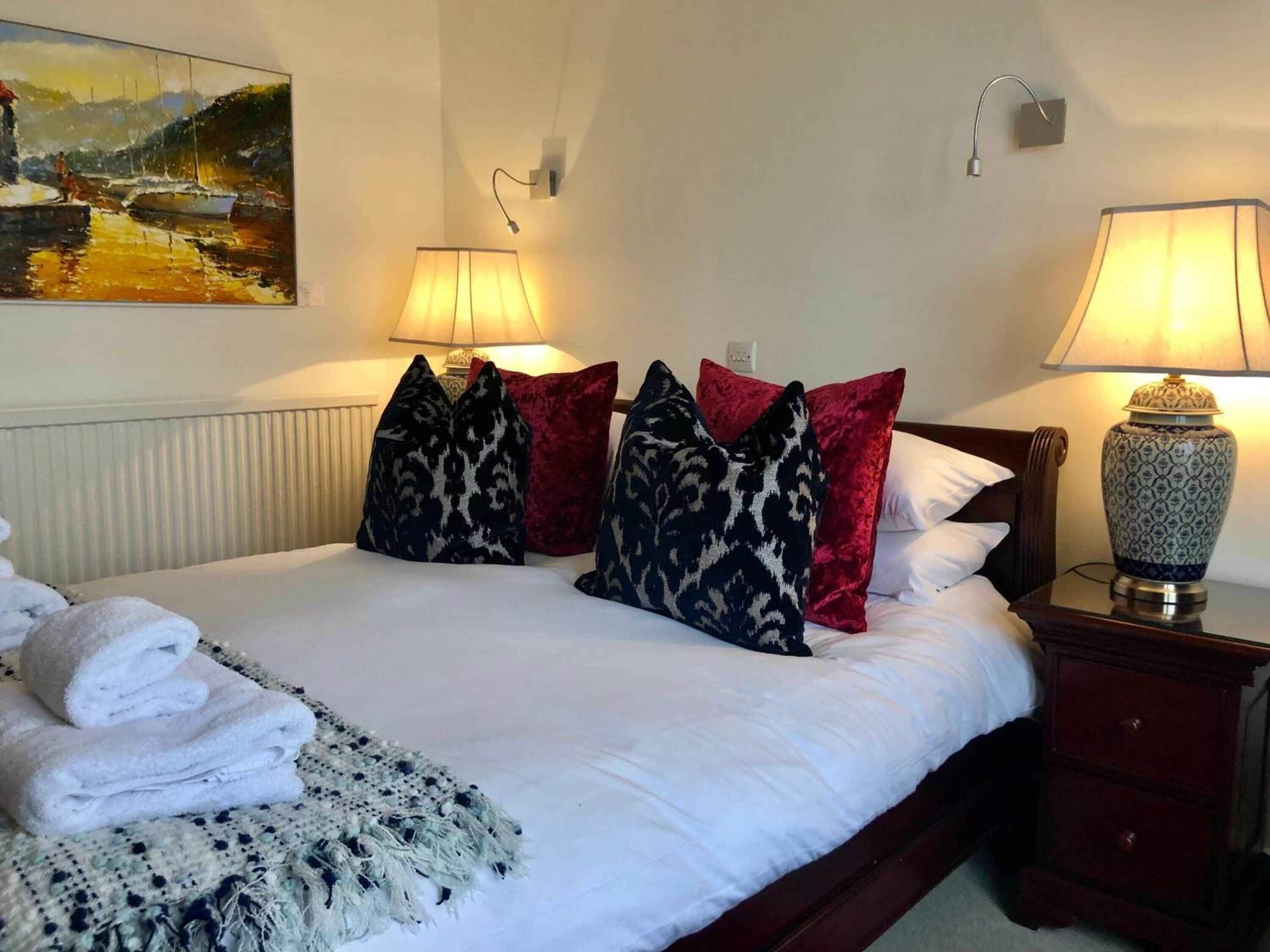 A neatly made bed with white linens, multiple pillows in various patterns, a folded towel, and a decorative lamp on a bedside table.