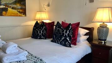 A neatly made bed with white linens, multiple pillows in various patterns, a folded towel, and a decorative lamp on a bedside table.