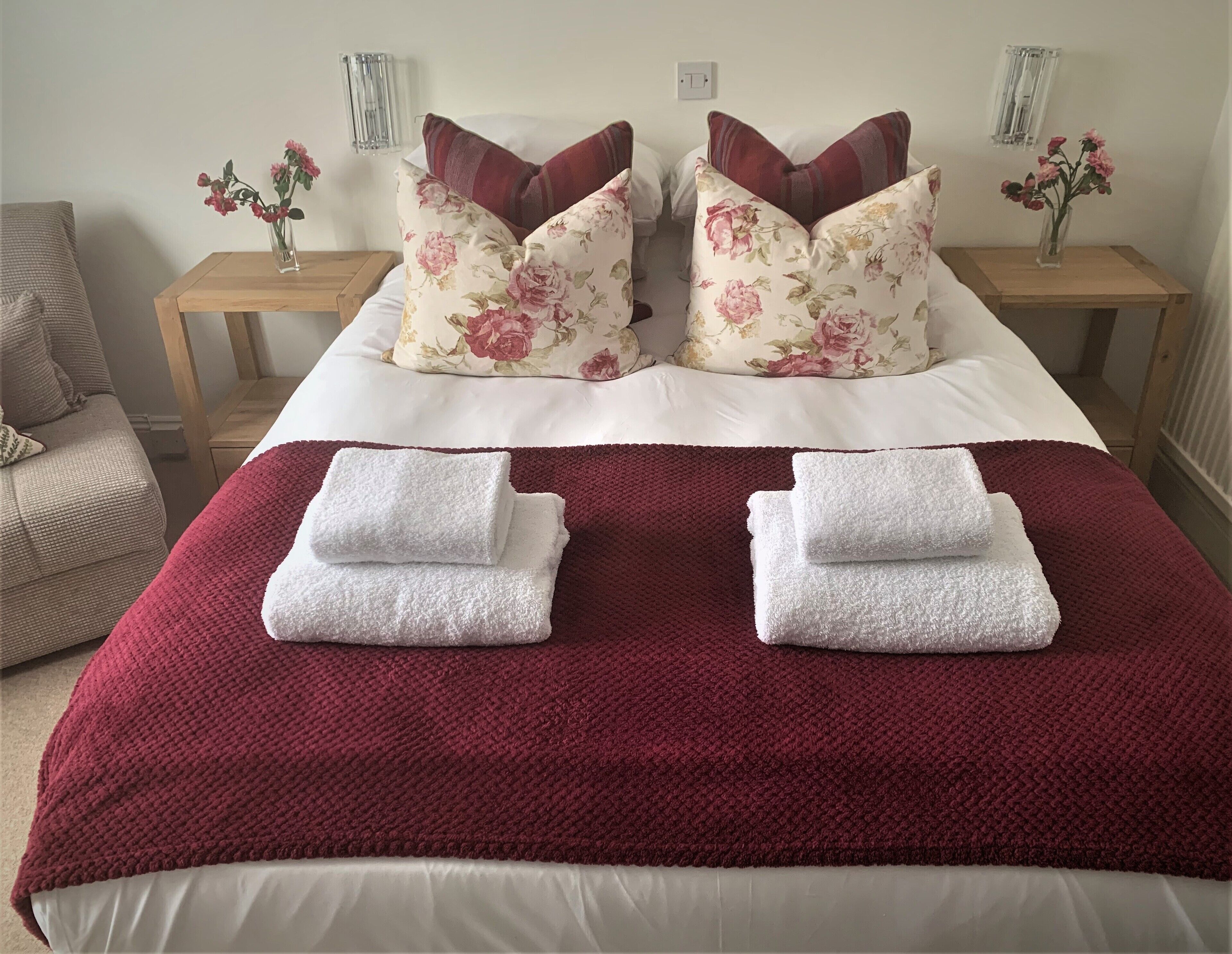 A neatly made bed with floral pillows, a red blanket, and two folded towels.
