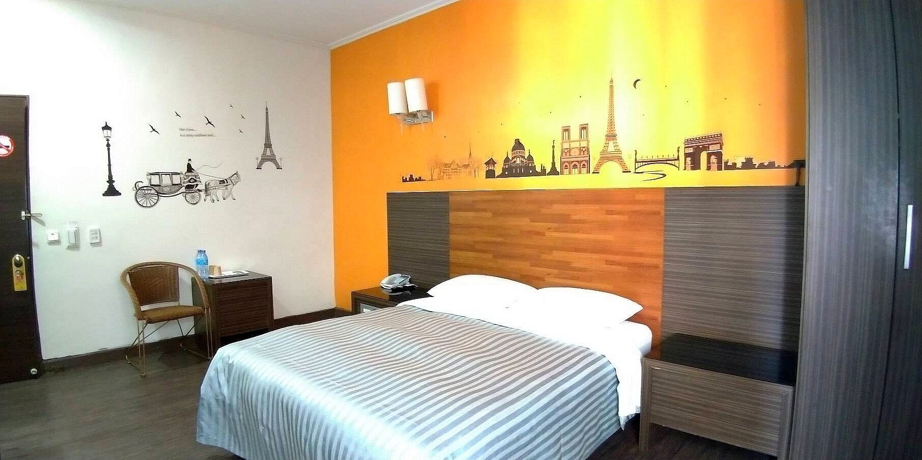A hotel room with a bed, a chair, a nightstand, and a wall decorated with cityscape silhouettes.