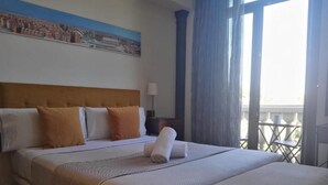 Hotellihuone, jossa on sänky, yöpöytä lampun kanssa, näkymä parvekkeelle ja taulu seinällä.