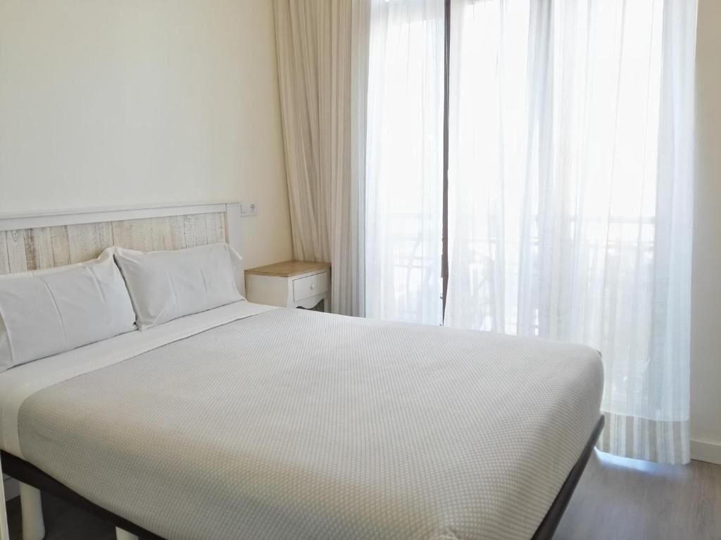 Una cama bien hecha con sábanas blancas en una habitación con cabecera de madera, una mesita de noche y cortinas traslúcidas.