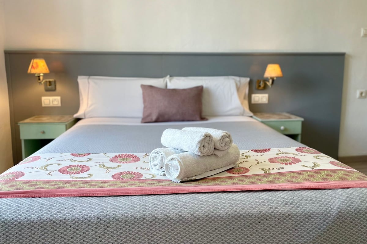 Una cama bien tendida con una colcha floral, dos almohadas y toallas enrolladas sobre una mesita de noche.