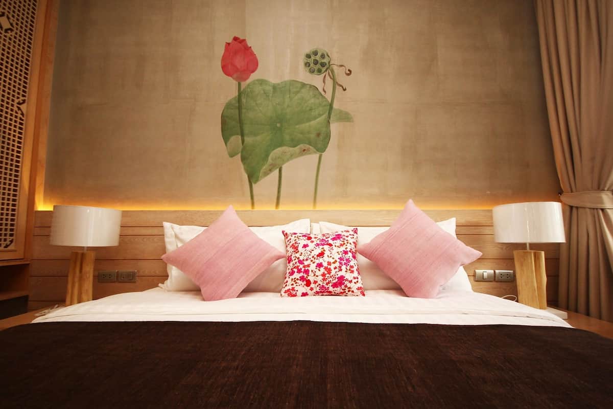 A hotel room with a bed, two bedside lamps, a floral painting on the wall, and a wooden headboard.