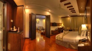 A hotel room with a wooden floor, a bed with a headboard, a desk with a lamp, and a bathroom with a bathtub and mirror. - Hotel Le Ruchi the Prince (Mysore)