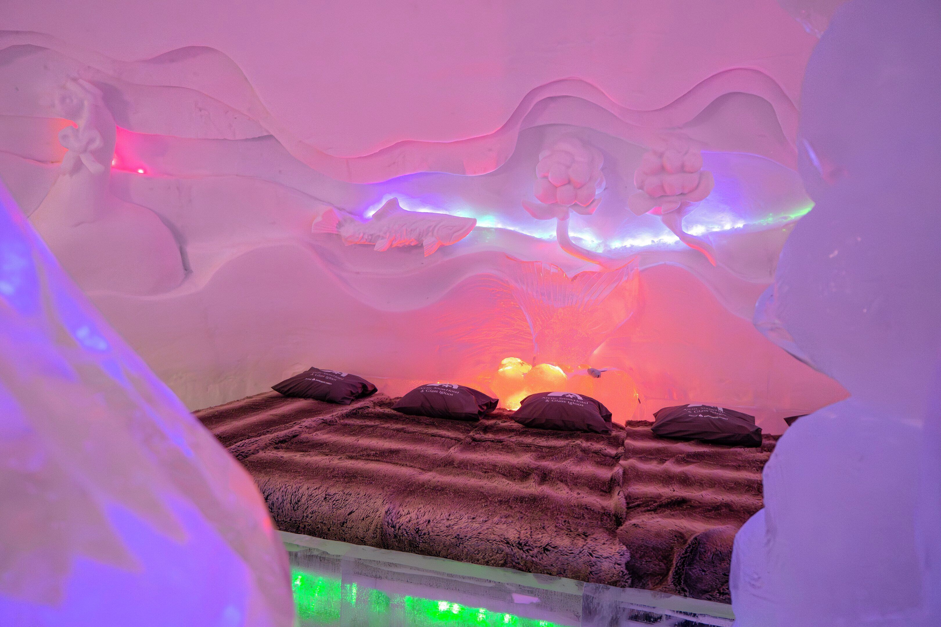An ice hotel room with a bed, pillows, and a unique ice sculpture.