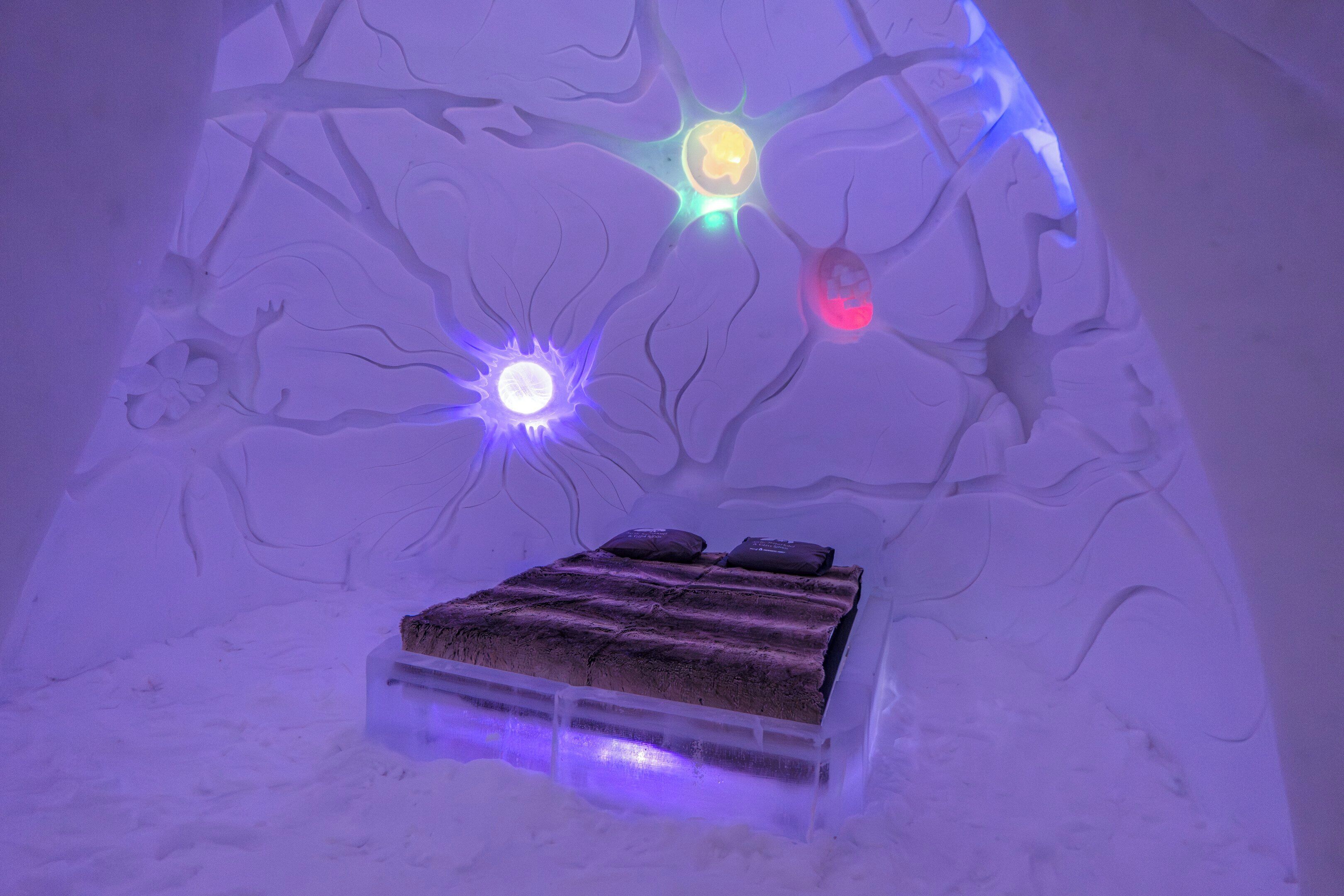 An ice hotel room with a bed, illuminated by colored lights and a tree-like ceiling design.