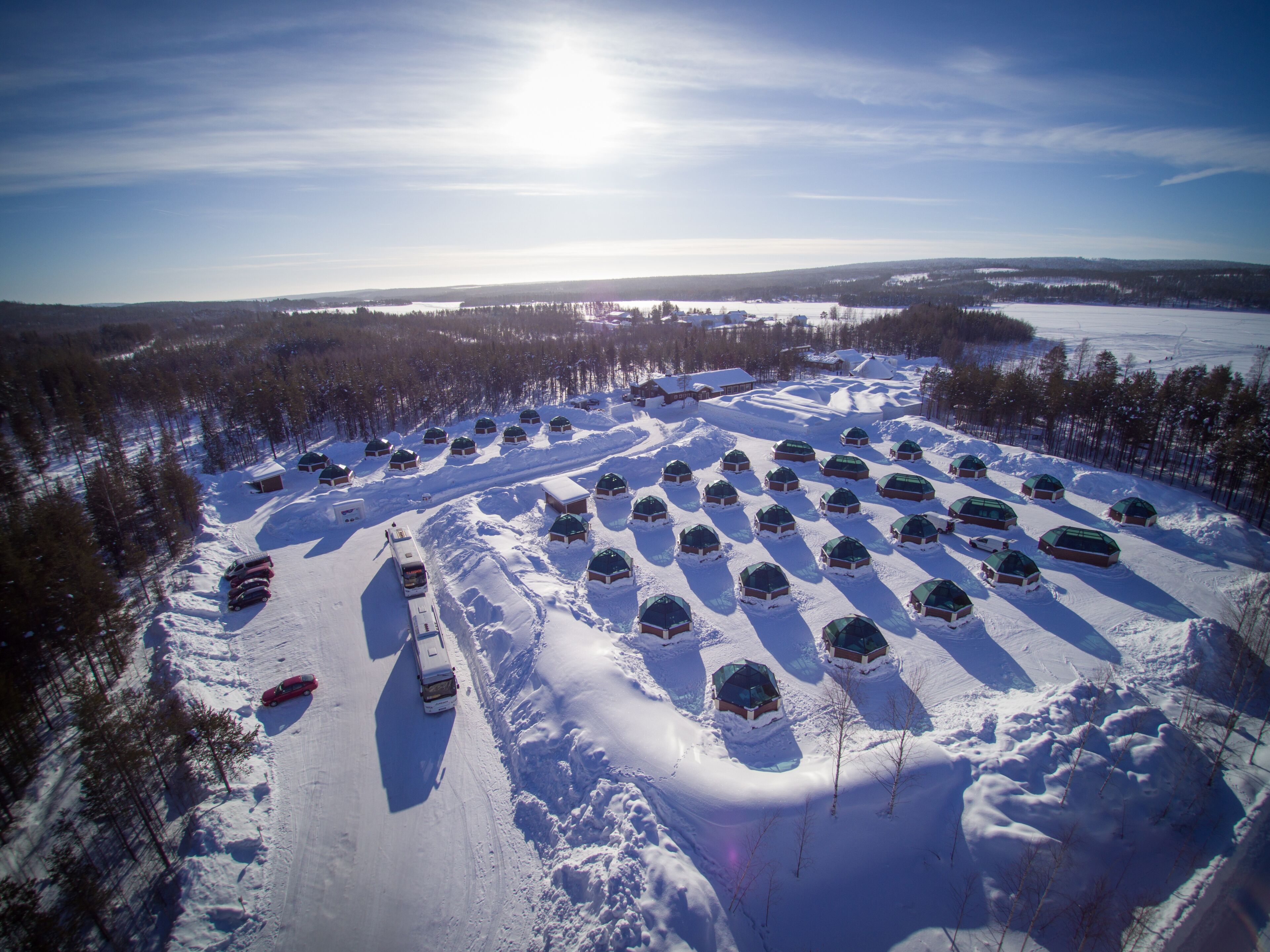 An aerial view of a snow-covered area with multiple dome-shaped cabins arranged in rows, a snowmobile parked, and a clear sky.