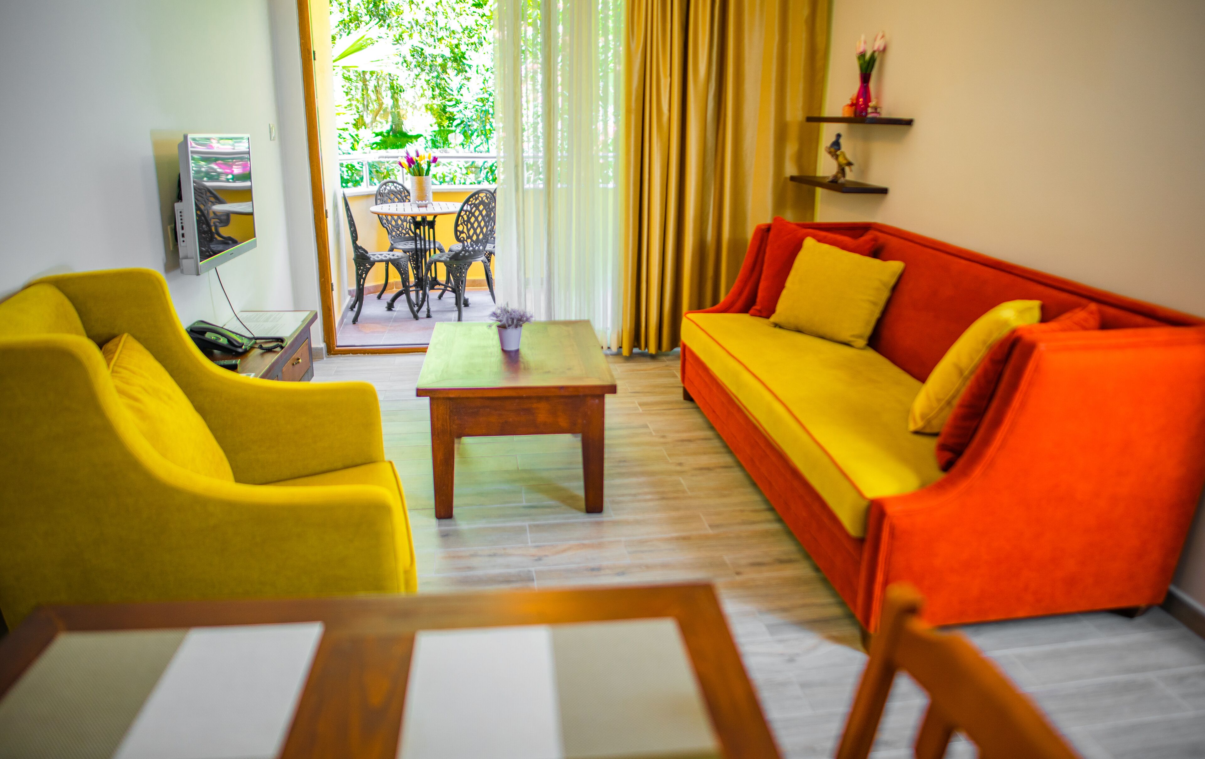 A living room with a yellow sofa, a red sofa, and a wooden coffee table. There is a dining table with chairs and a window with curtains.
