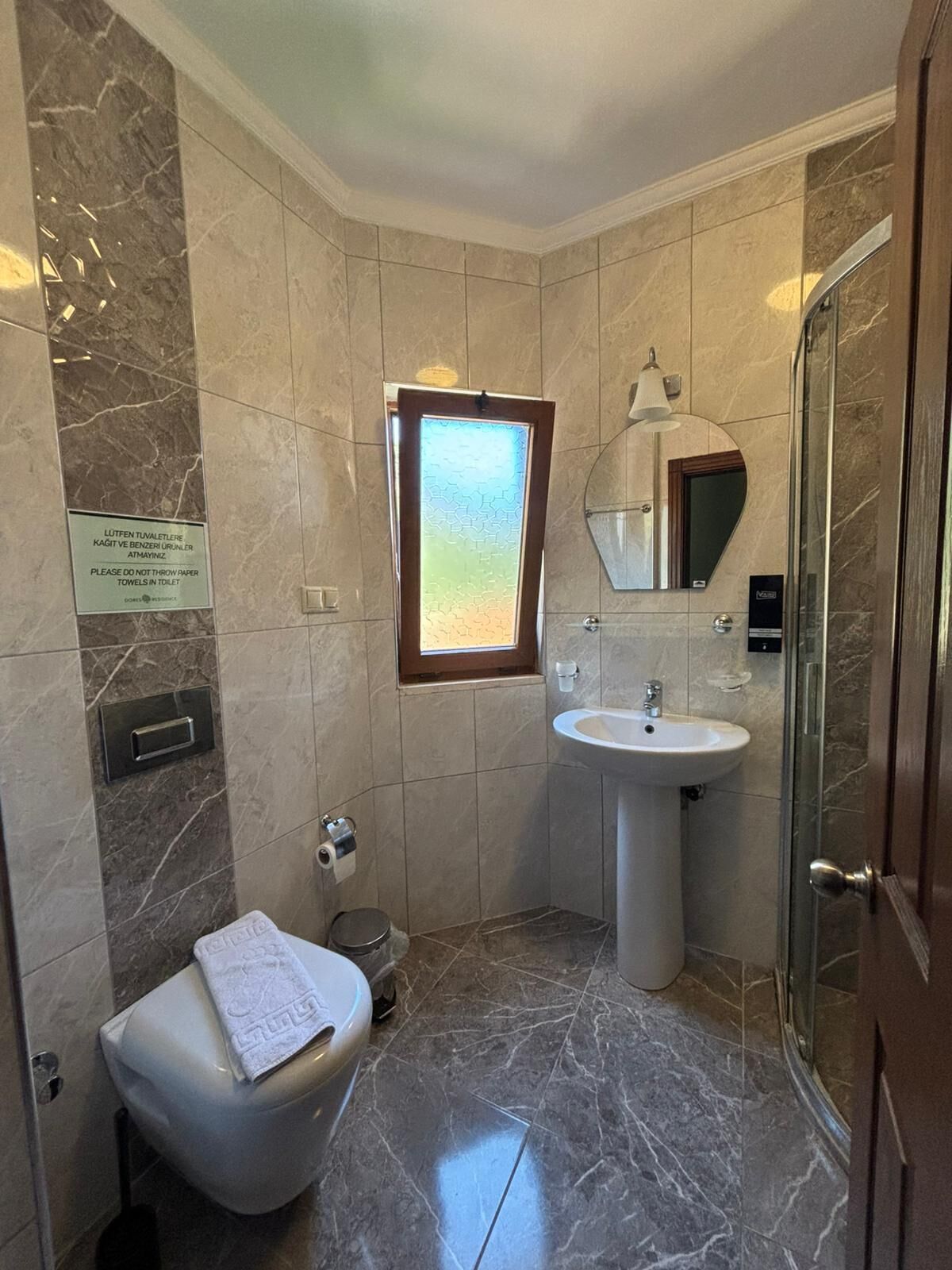 Een moderne badkamer met toilet, wastafel en douche. Een bordje geeft aan dat er geen paperhanddoeken in het toilet mogen worden gegooid.