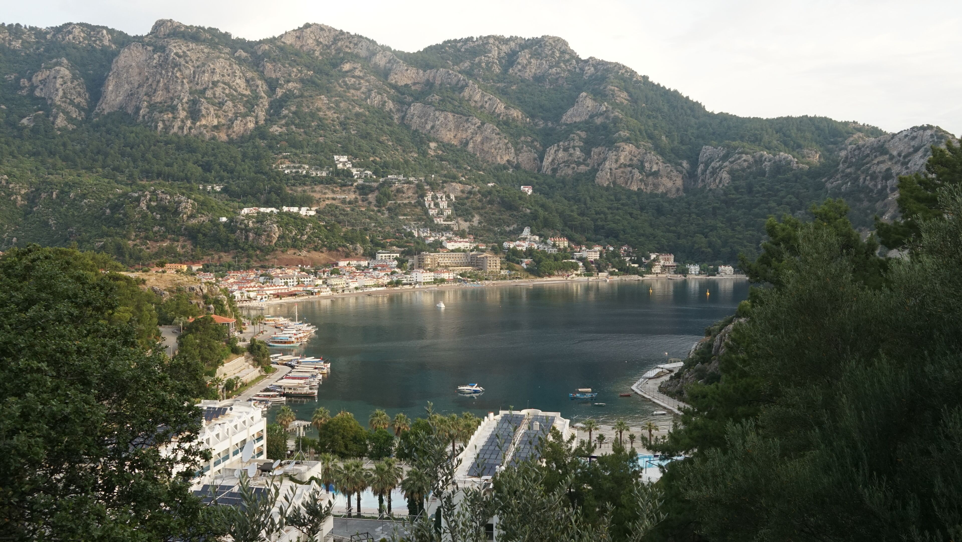 A coastal town with a marina, buildings, and mountains in the background.