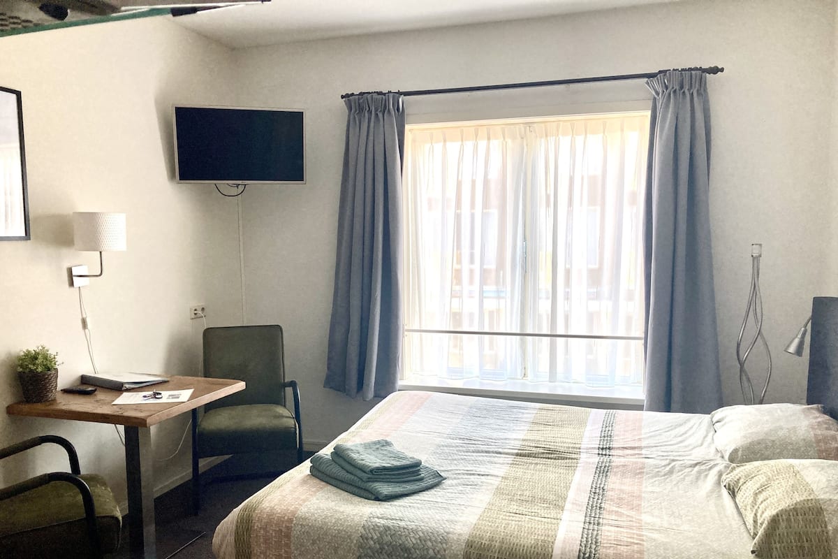 Quarto de hotel com cama, escrivaninha, cadeira, TV fixada na parede e janela com cortinas.