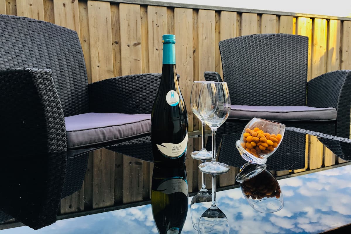 Uma garrafa de vinho, duas taças e uma tigela de petiscos sobre uma mesa de vidro ao ar livre.