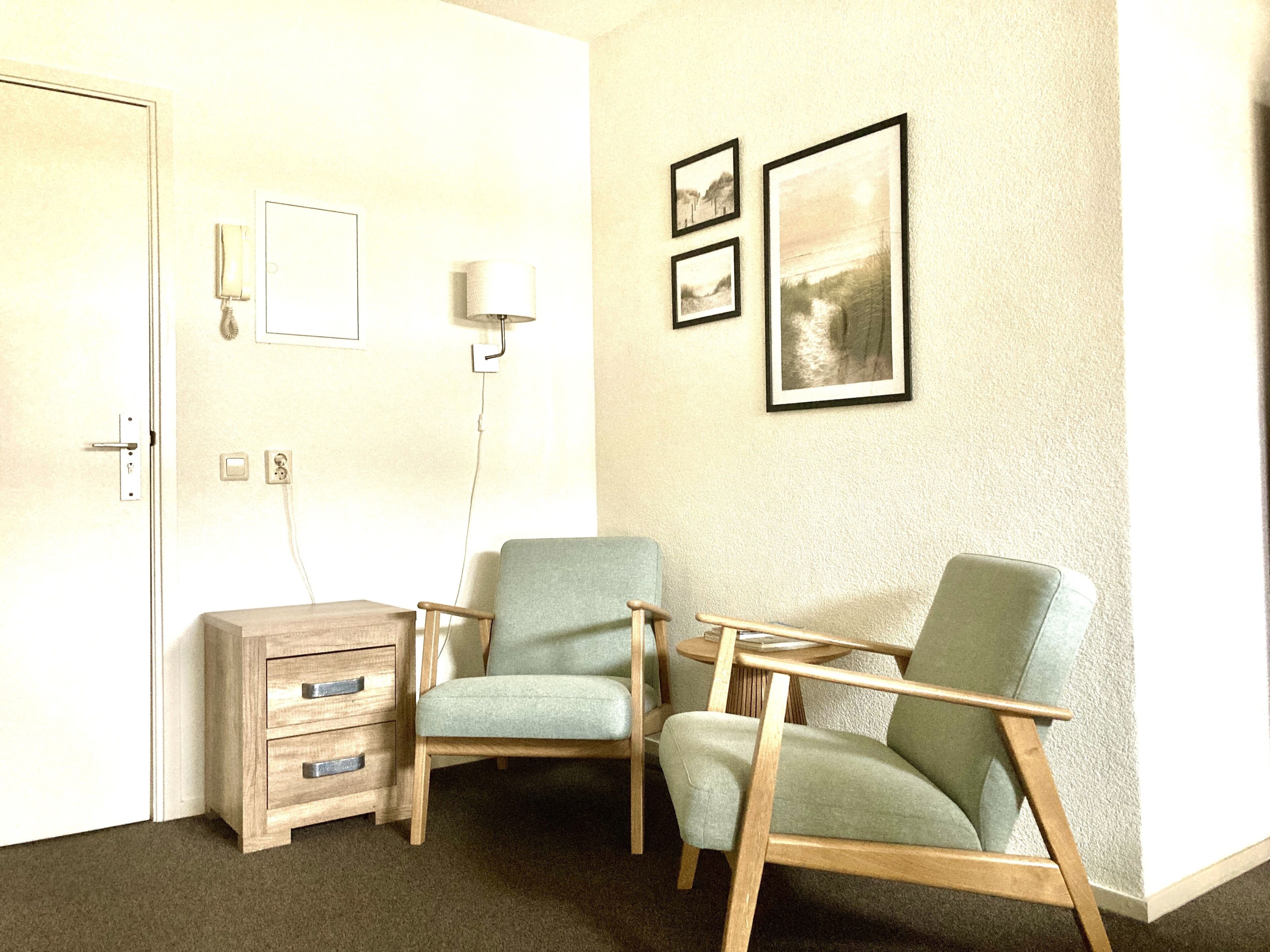 A room with two green armchairs, a wooden side table, a small wooden dresser, and a wall-mounted lamp. There are two framed pictures on the wall.