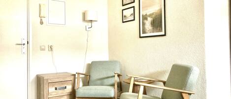 A room with two green armchairs, a wooden side table, a small wooden dresser, and a wall-mounted lamp. There are two framed pictures on the wall.