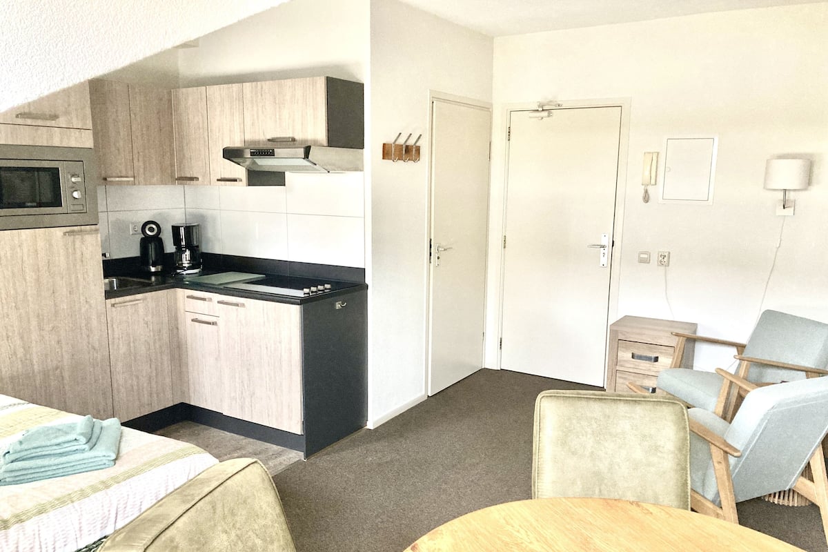 Quarto de hotel compacto com cozinha americana, uma cama, uma mesa de madeira redonda e duas poltronas.