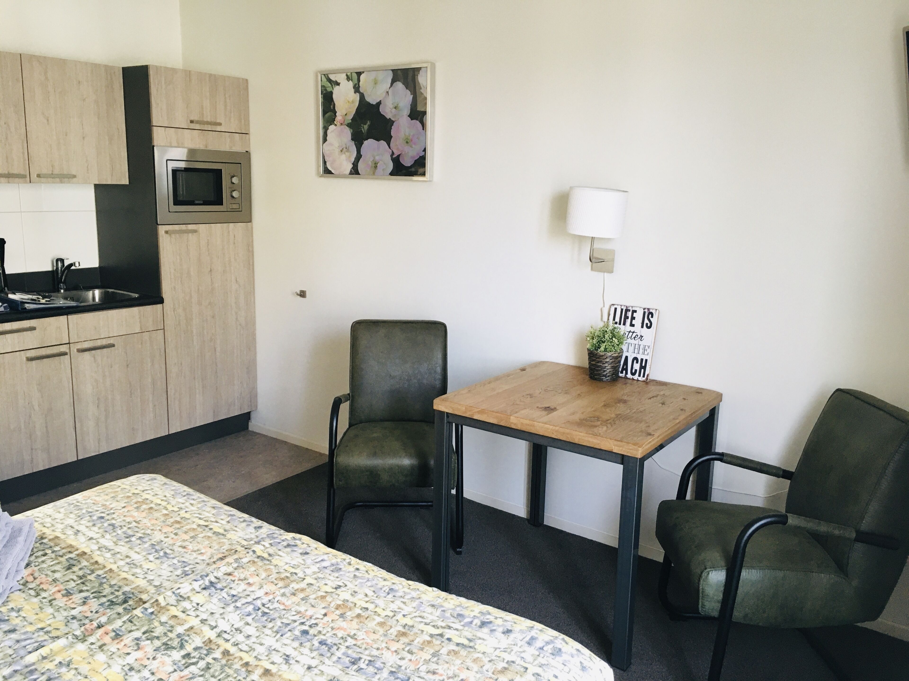 A compact hotel room with a bed, two chairs, a small table, a microwave, a sink, and a wall-mounted lamp.