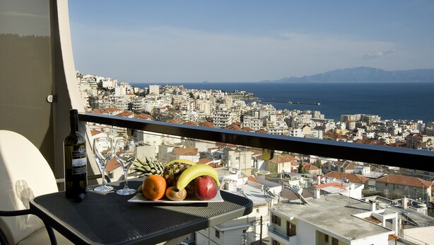 A balcony with a table holding a bottle of wine, two glasses, and a plate with fruits. The view includes a coastal cityscape and the sea. - Egnatia City Hotel & Spa (Kavala)