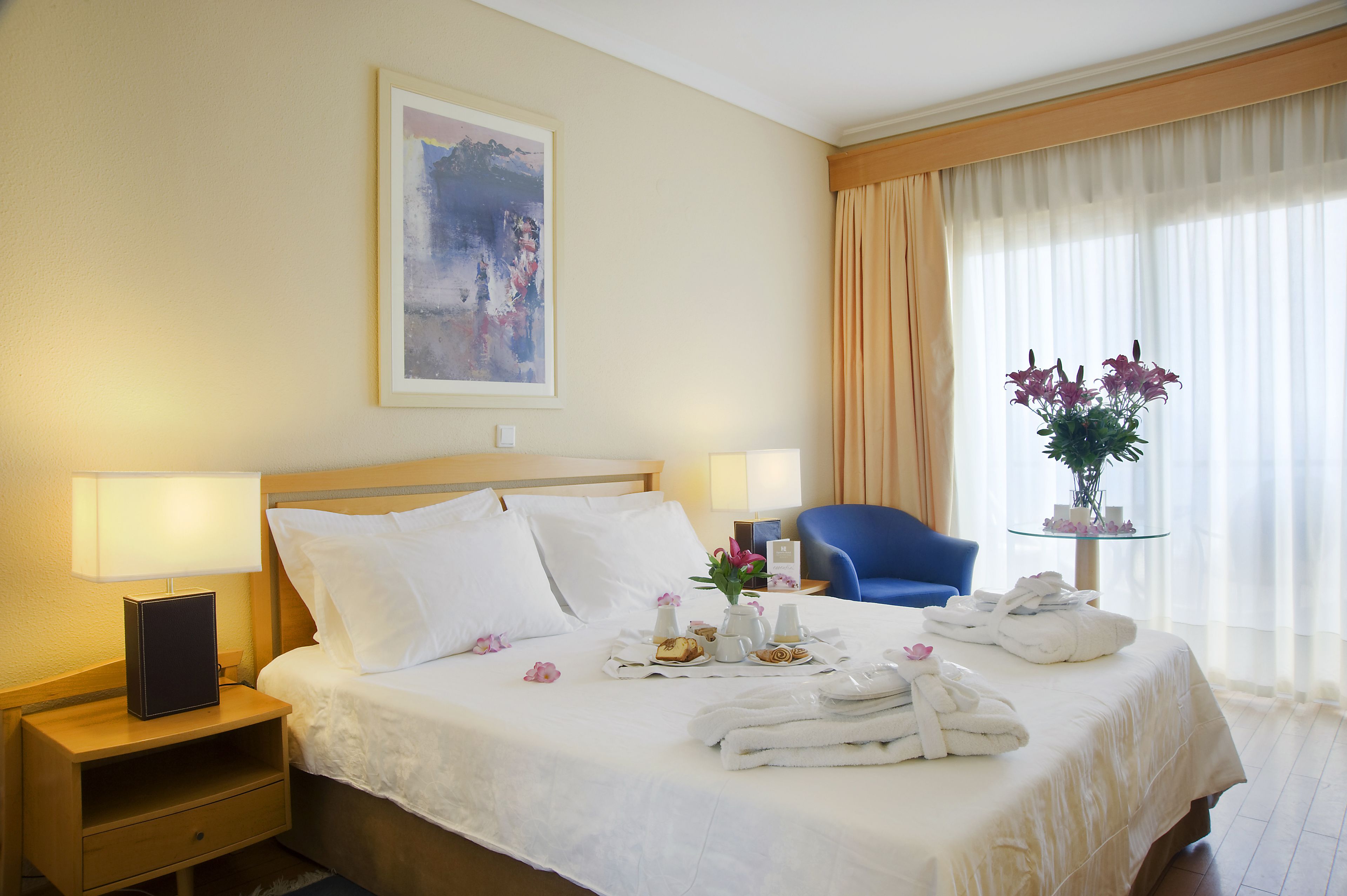 Un cuarto de hotel con una cama bien hecha, dos mesitas de noche con lámparas, una silla, una pequeña mesa con un jarrón de flores y un cuadro en la pared.