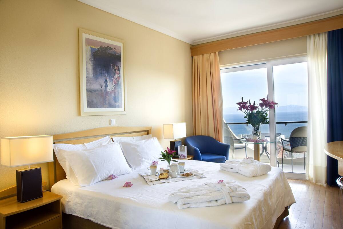 Ein Hotelzimmer mit Bett, Nachttisch, Tisch mit Blumenvase und Blick auf die Stadt durch große Fenster.