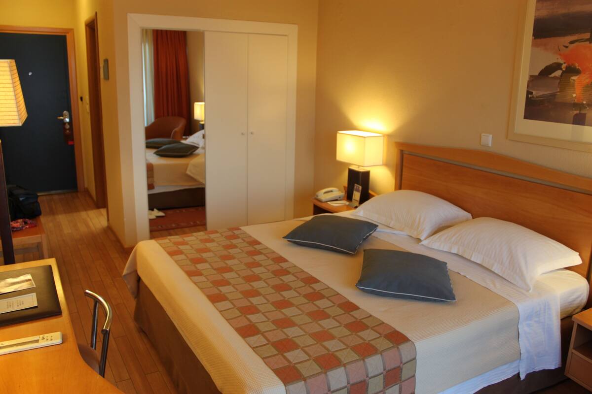 Ein Hotelzimmer mit einem Bett, einem Nachttisch, einer Lampe, einem Sessel, einem Schreibtisch und einer Tür zu einem anderen Raum.