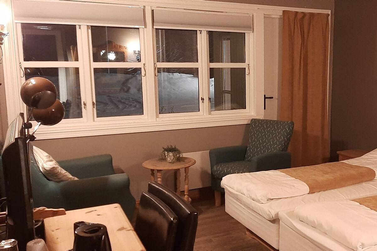 Quarto de hotel com duas camas, uma mesa pequena, uma cadeira, um abajur e uma janela com cortinas.