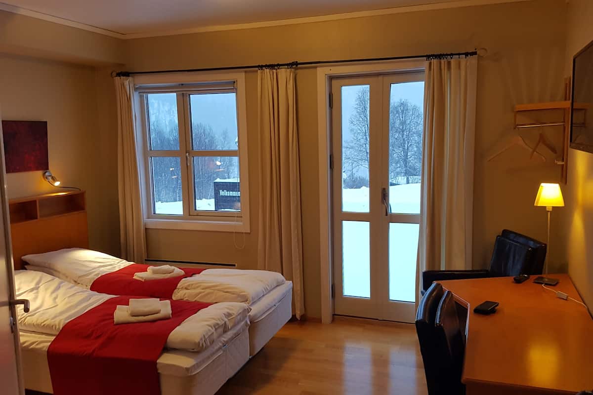 Quarto de hotel com cama, escrivaninha, cadeira e janela com cortinas.