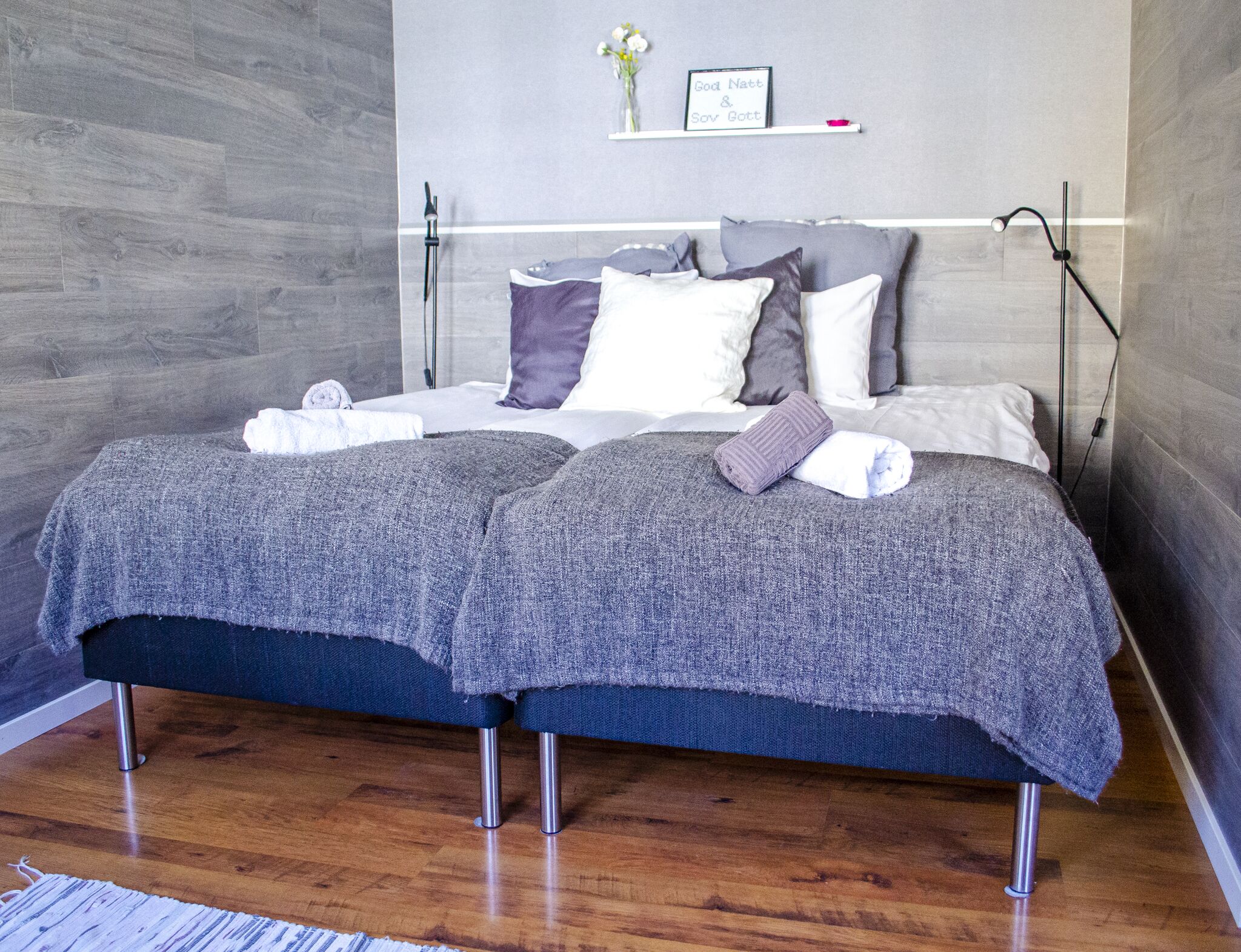 Uma cama de casal com roupa de cama cinzenta, uma mesa de cabeceira e uma prateleira com um objeto emoldurado.