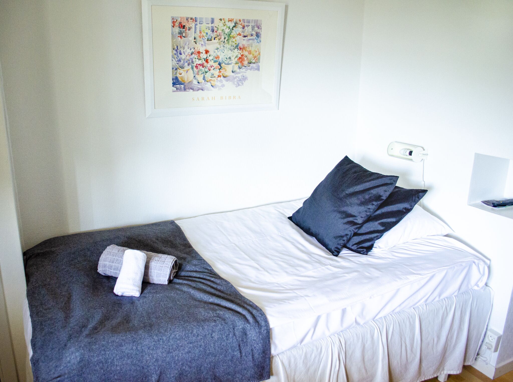 A single bed with white linens, a dark blue pillow, and a grey blanket. A framed artwork hangs on the wall above the bed.