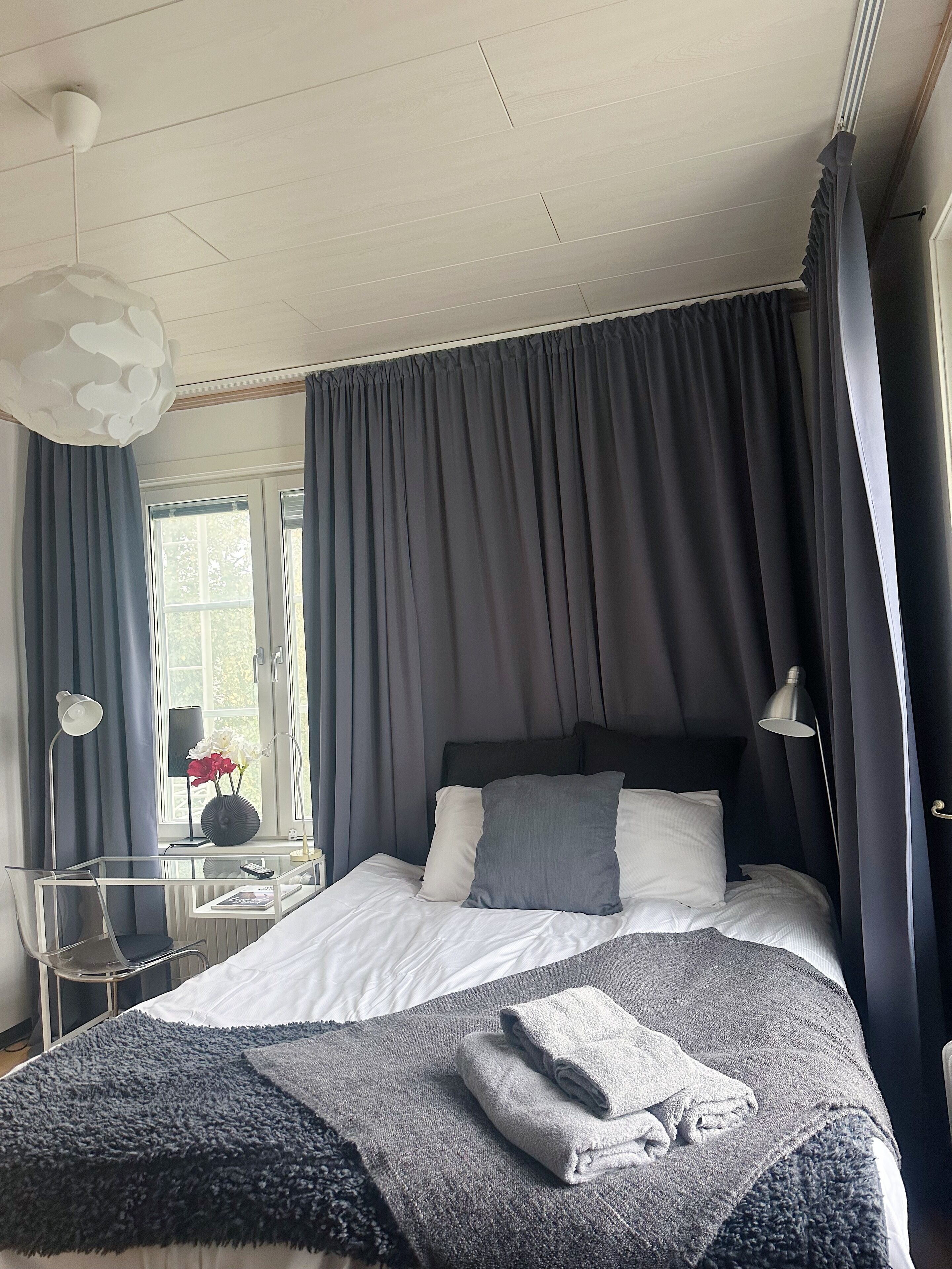 A modern bedroom with a large bed, dark curtains, a bedside table with a lamp, and a window with a view of greenery.