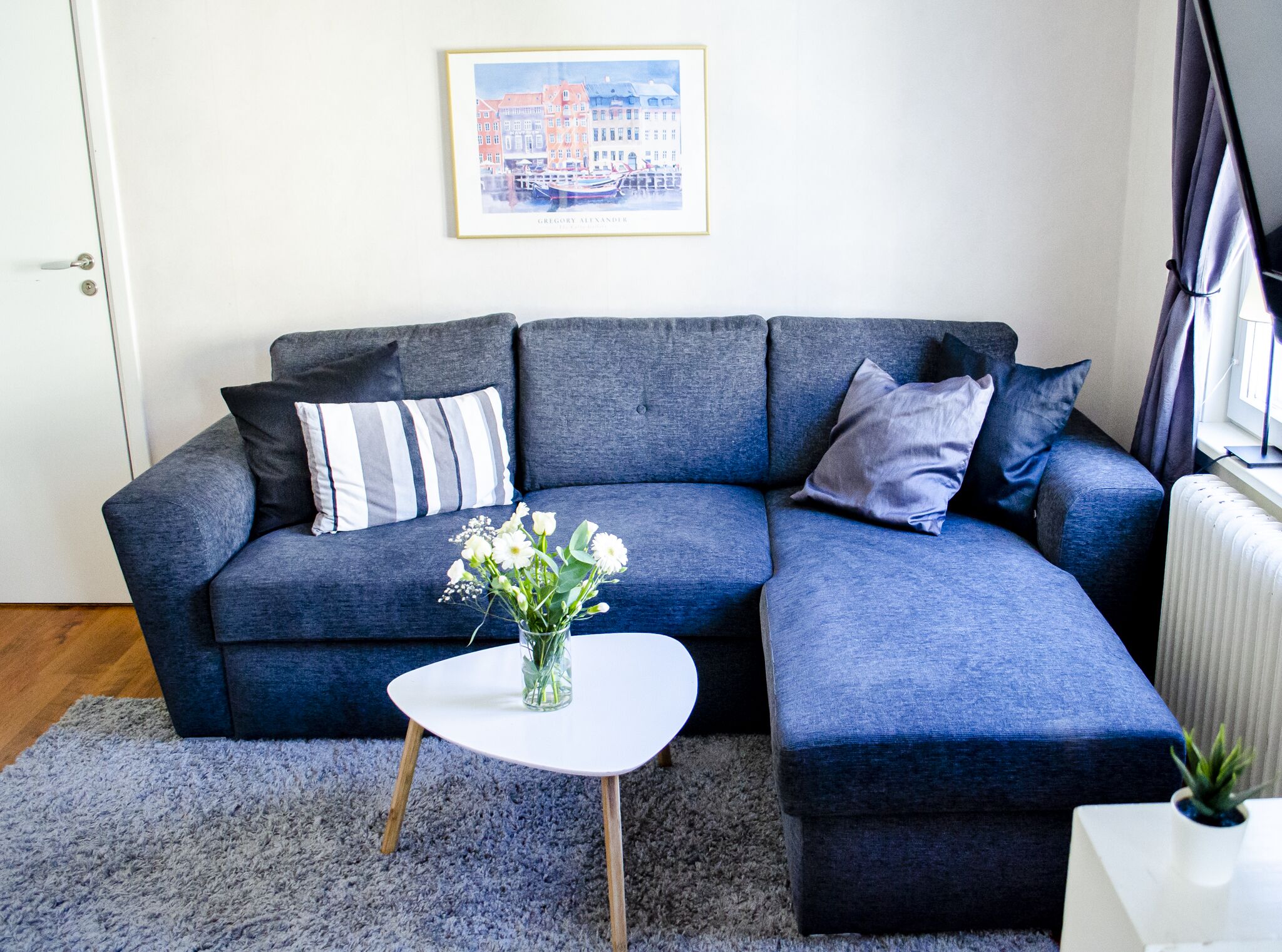 A modern living room with a grey sectional sofa, a small white side table, and a framed cityscape picture on the wall.