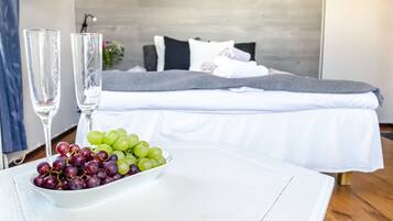A room with a bed, a bedside table with two glasses and a bowl of grapes, and a grey blanket on the bed.