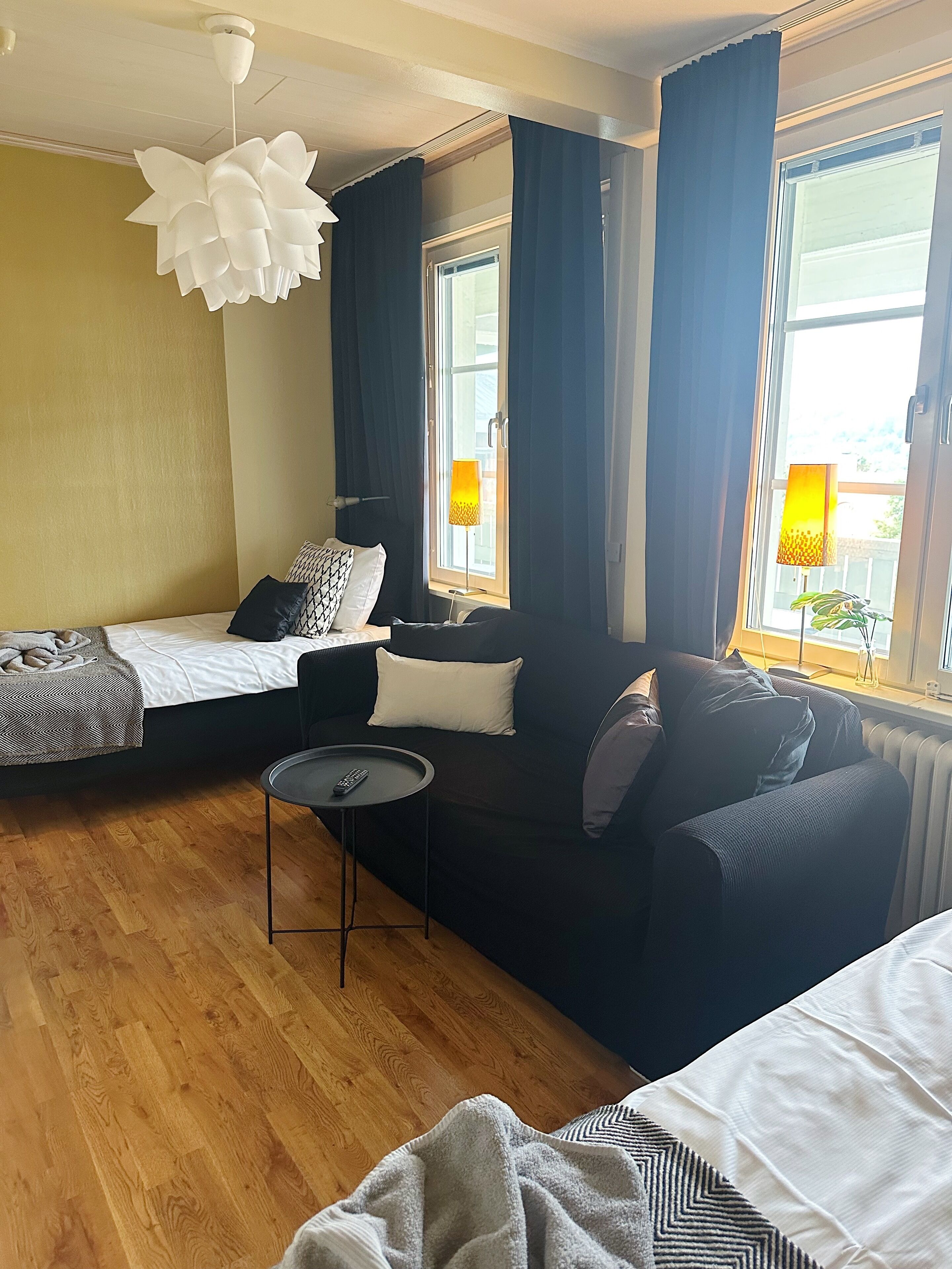 Een hotelkamer met een bed, een slaapbank, een kleine tafel met een afstandsbediening en een raam met gordijnen.