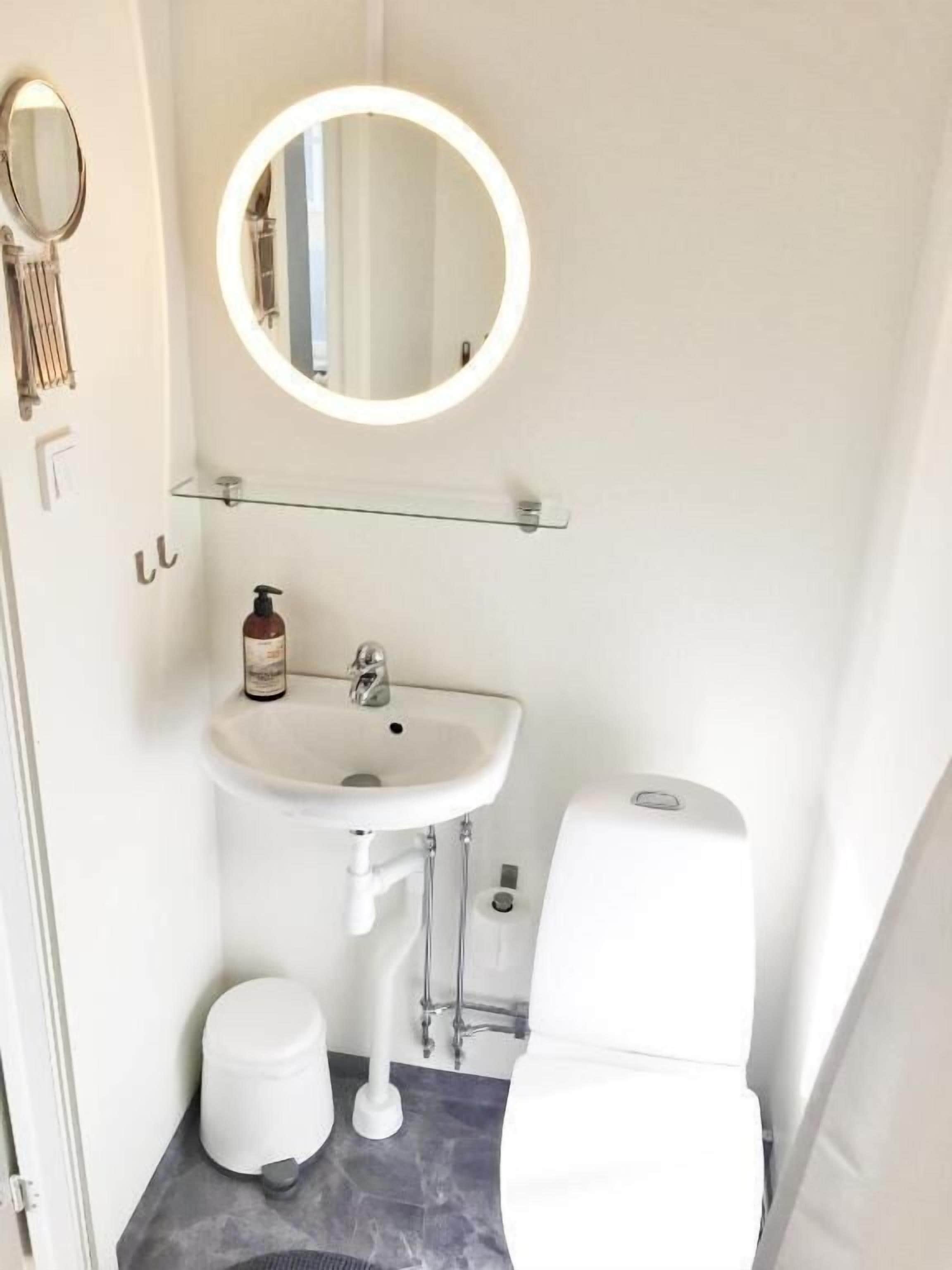 A small, modern bathroom with a white sink, a round mirror, a toilet, and a small trash bin.