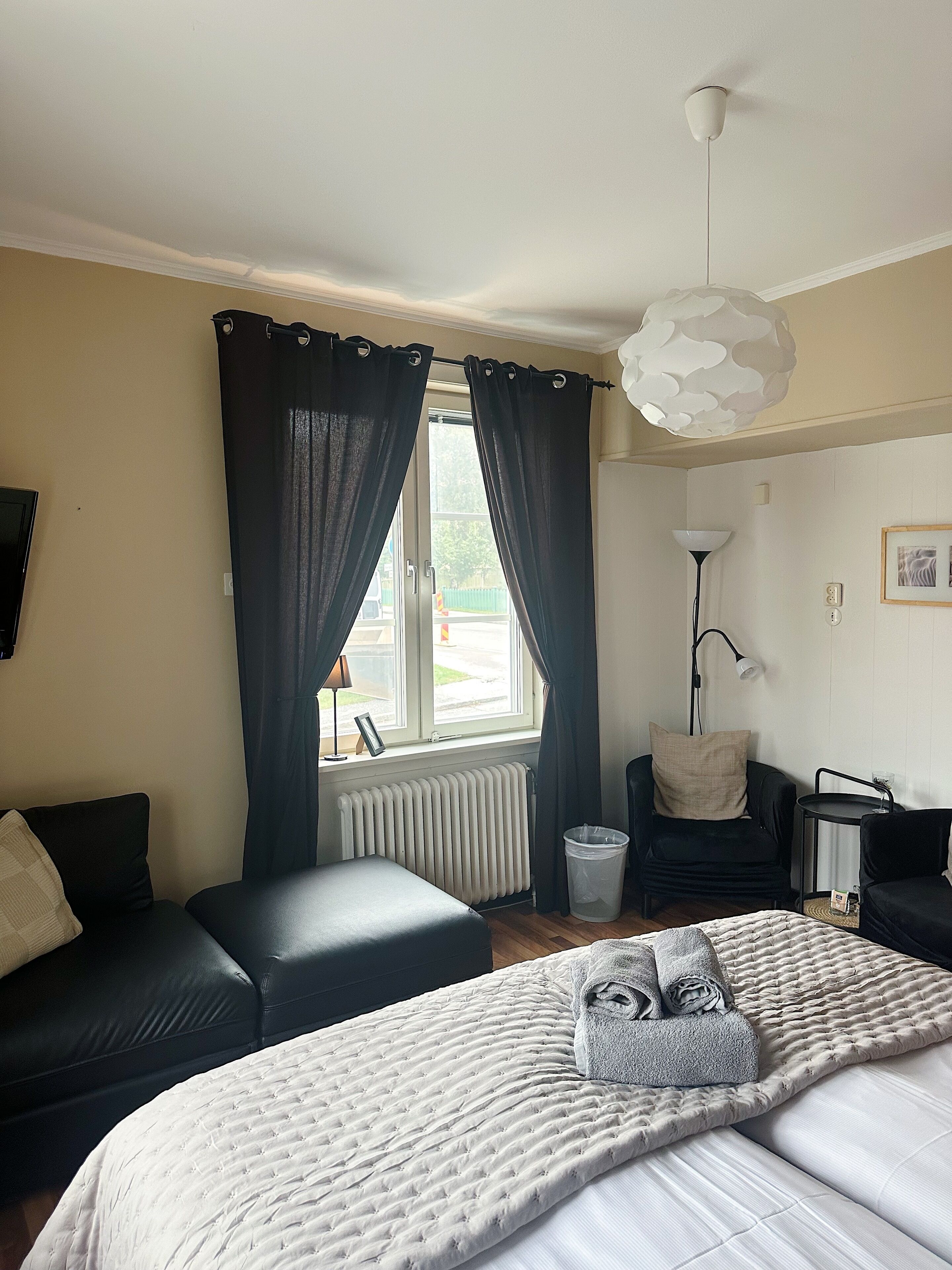 Quarto com cama, sofá, poltrona, luminária de chão e janela com cortinas.