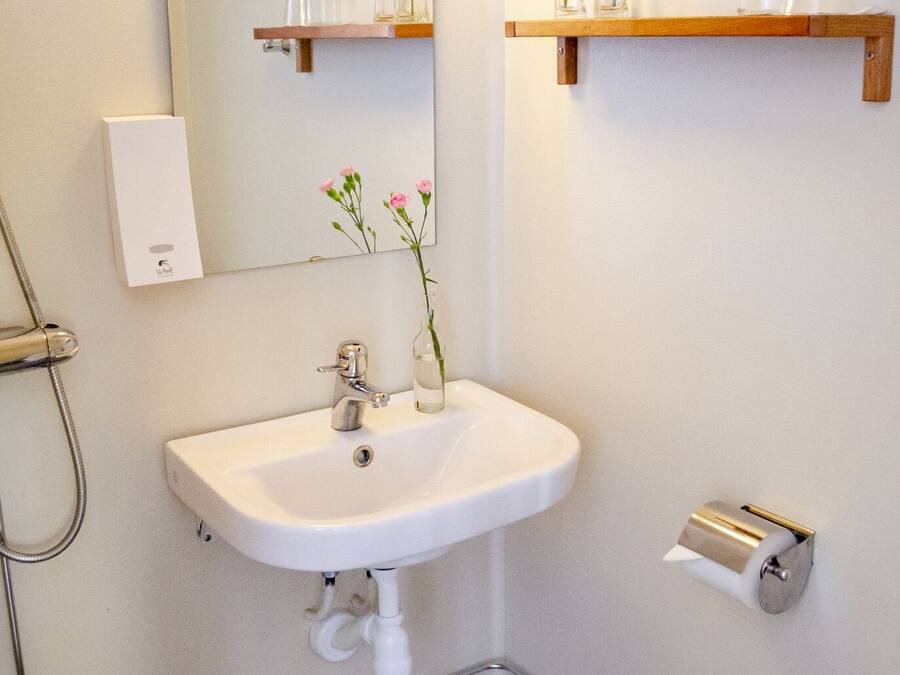 A bathroom with a white sink, a mirror, a towel dispenser, a toilet paper holder, and a shelf with vases.