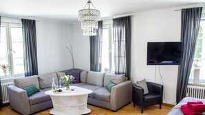 Una sala de estar moderna con un sofá modular, una mesa de centro redonda, un sillón negro, un televisor de pantalla plana y una cama con ropa de cama roja.