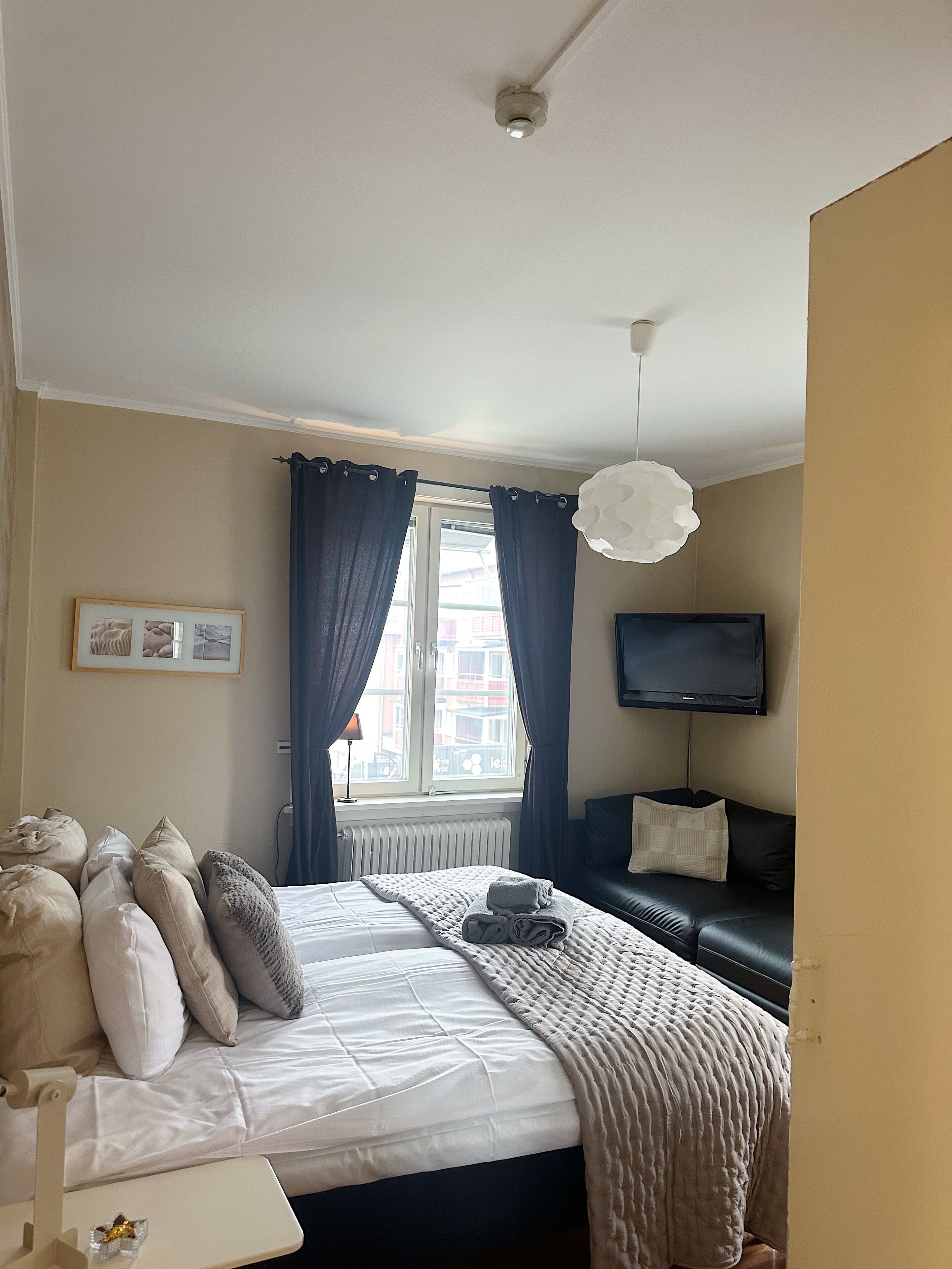 Una camera da letto con un letto, un comodino, una lampada, un televisore, una finestra con tende e vista sugli edifici circostanti.