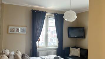Una camera da letto con un letto, un comodino, una lampada, un televisore, una finestra con tende e vista sugli edifici circostanti.
