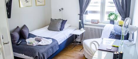 A compact hotel room with a bed, bedside table, lamp, and a view of the street through a window.