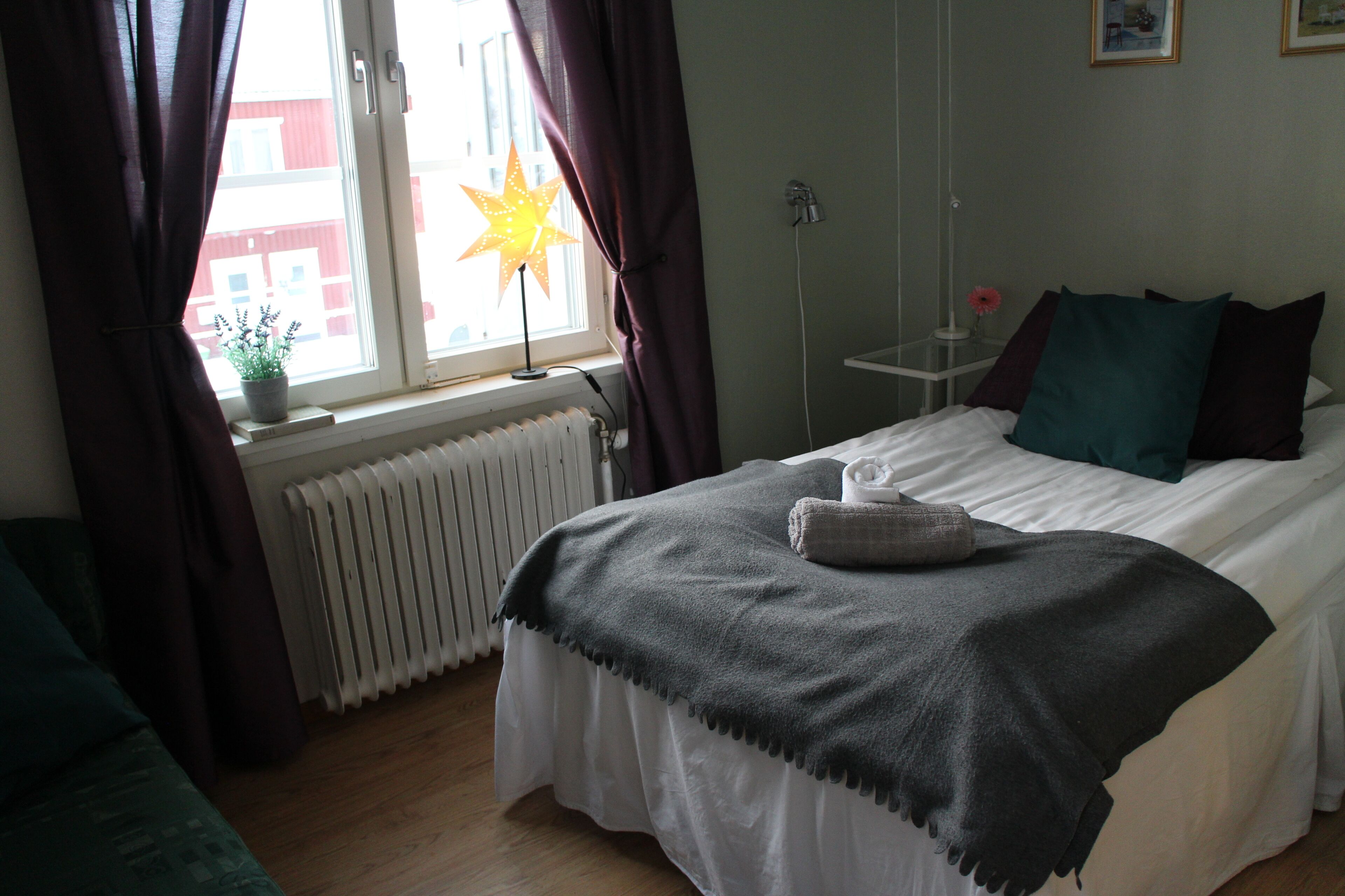 A bedroom with a bed, a radiator, a window with curtains, a star-shaped lamp, and a small table with a flower.