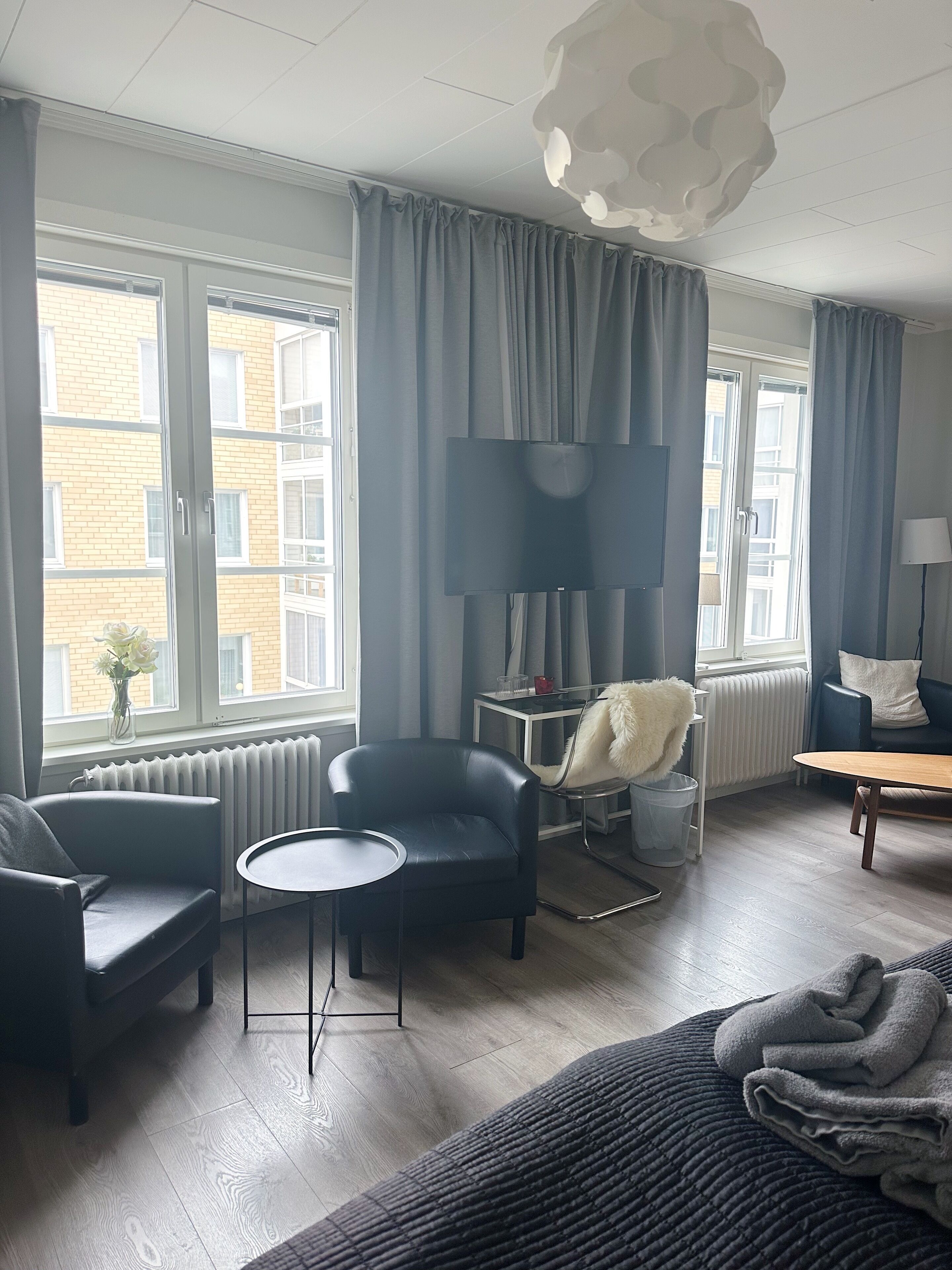 A modern living room with a sofa, armchair, and a round coffee table. There is a large window with curtains, a radiator, and a lamp on a side table.